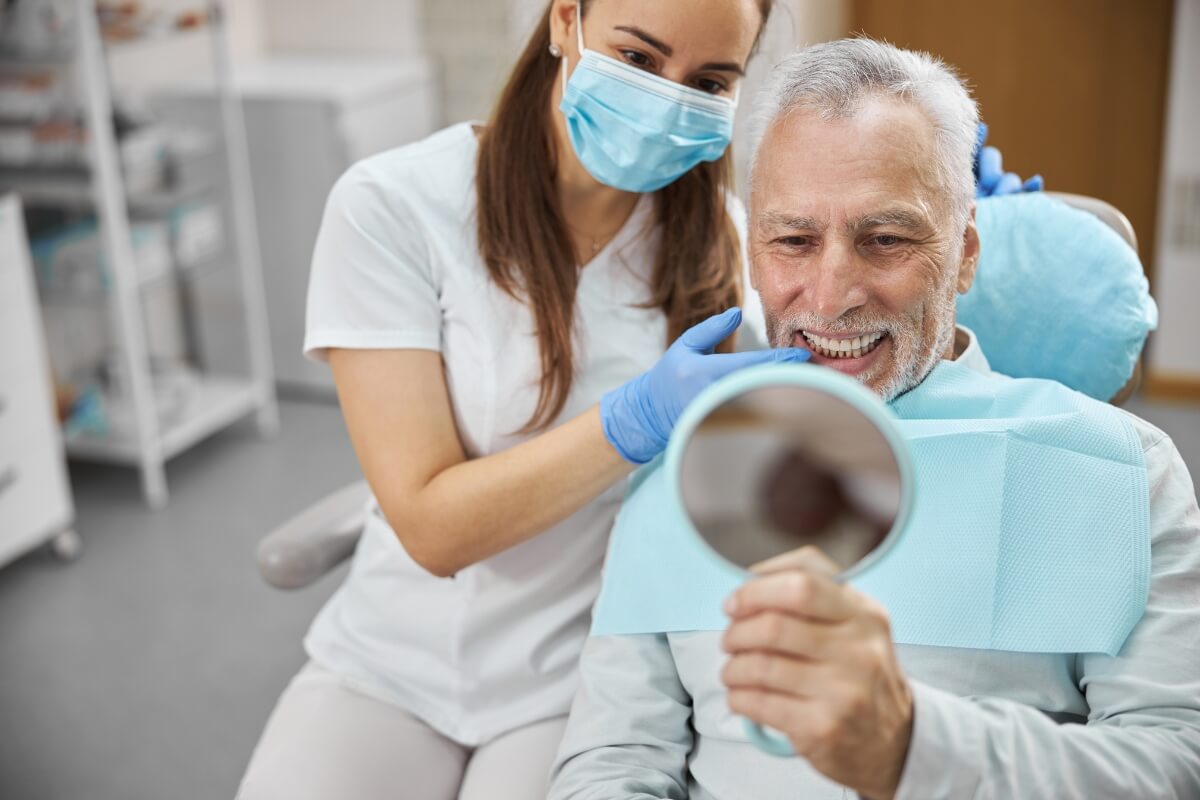 Old-Man-Smiling-While-Seeing-His-Teeth-In Mirror Old Man Smiling While Seeing His Restored Teeth In The Mirror