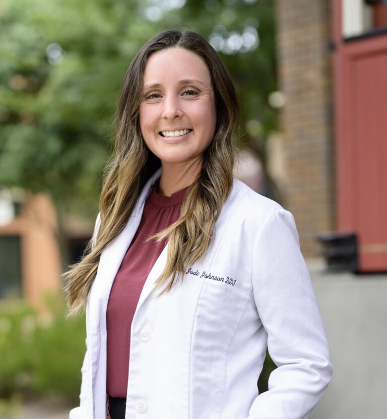 Dr. Jade Johnson stands outdoors in a white coat, smiling warmly in a professional setting at Dental Visions.
