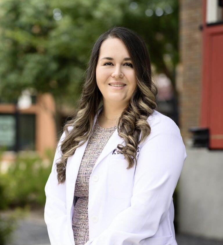 Dr. Kristina Chandler stands outdoors in a white coat, smiling warmly in front of Dental Visions, presenting a confident and approachable professional appearance.