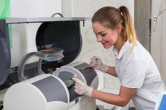 Person in white polo and gloves operates dental milling machine, holding crown material near transparent cover with rotating components.